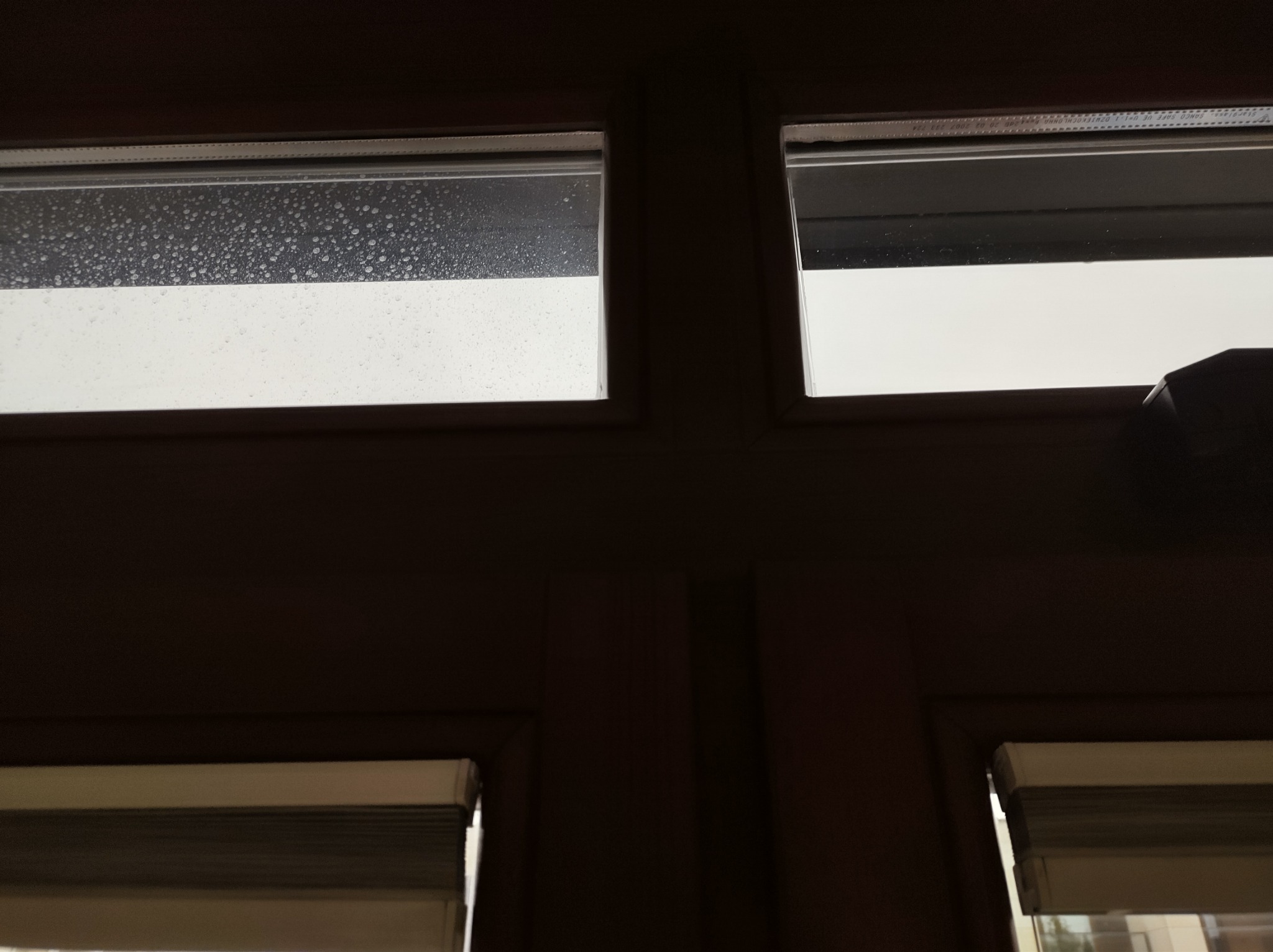 Okno z drewnianą ramą, górna lewa szyba pokryta kroplami wody, pozostałe czyste, dolne okna zasłonięte roletami.