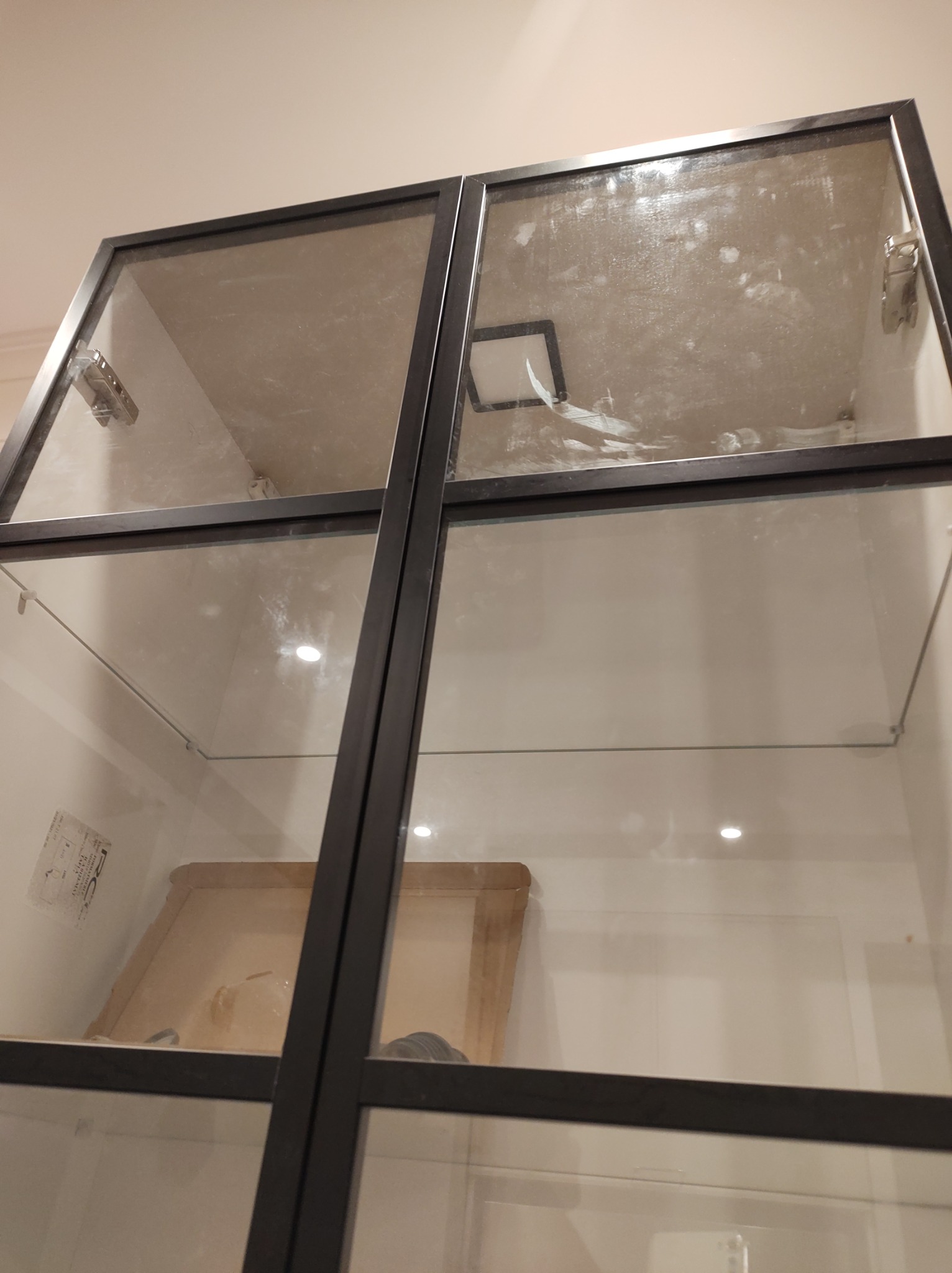 Wnętrze szklanej witryny z metalową ramą, widoczne ślady kurzu i zabrudzeń na szybach oraz półkach, oświetlenie punktowe wewnątrz.