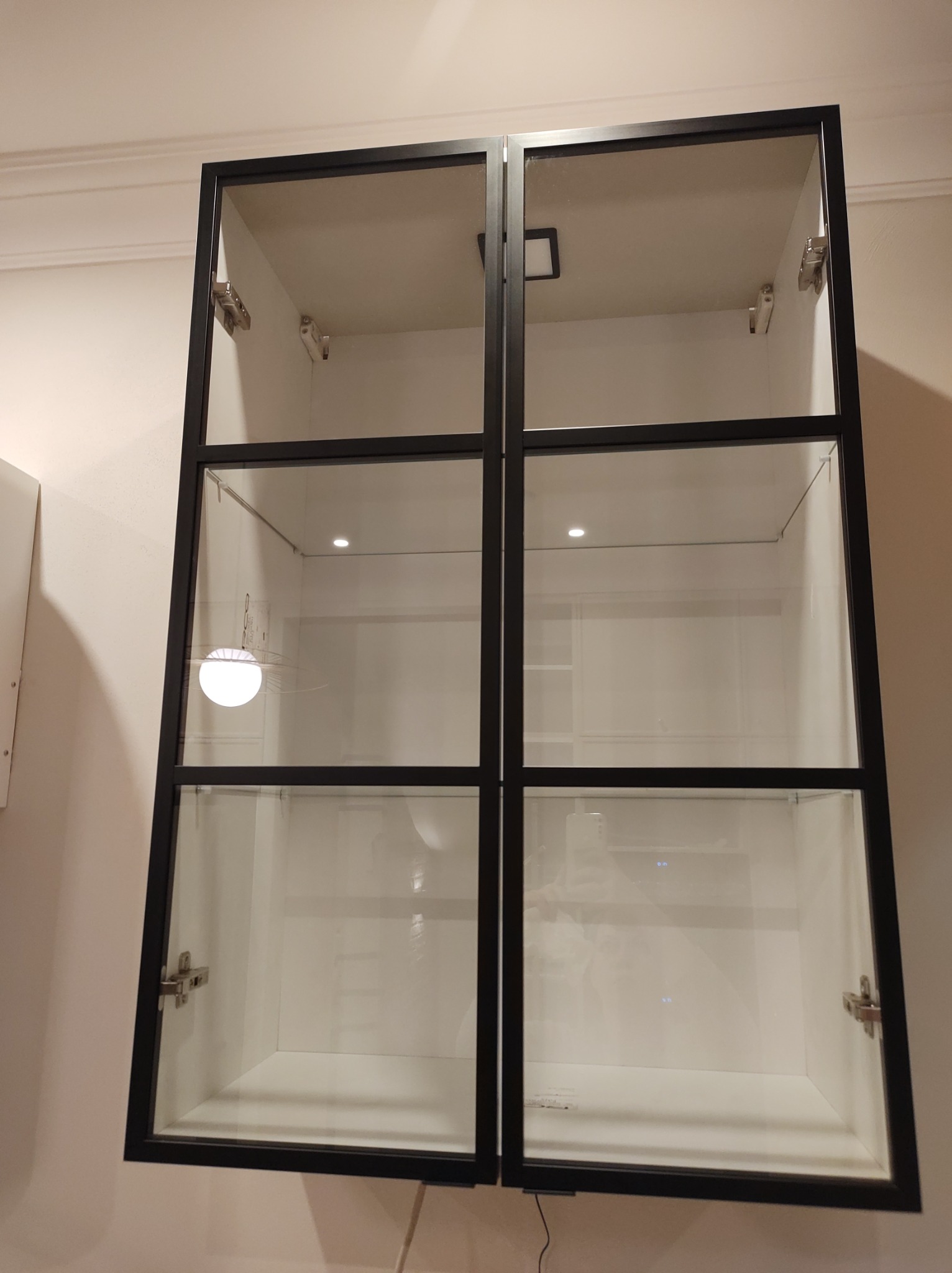 Pusta, czysta, biała szafka wisząca z przeszklonymi drzwiami w czarnej ramie, oświetlona od wewnątrz, zamontowana na ścianie.