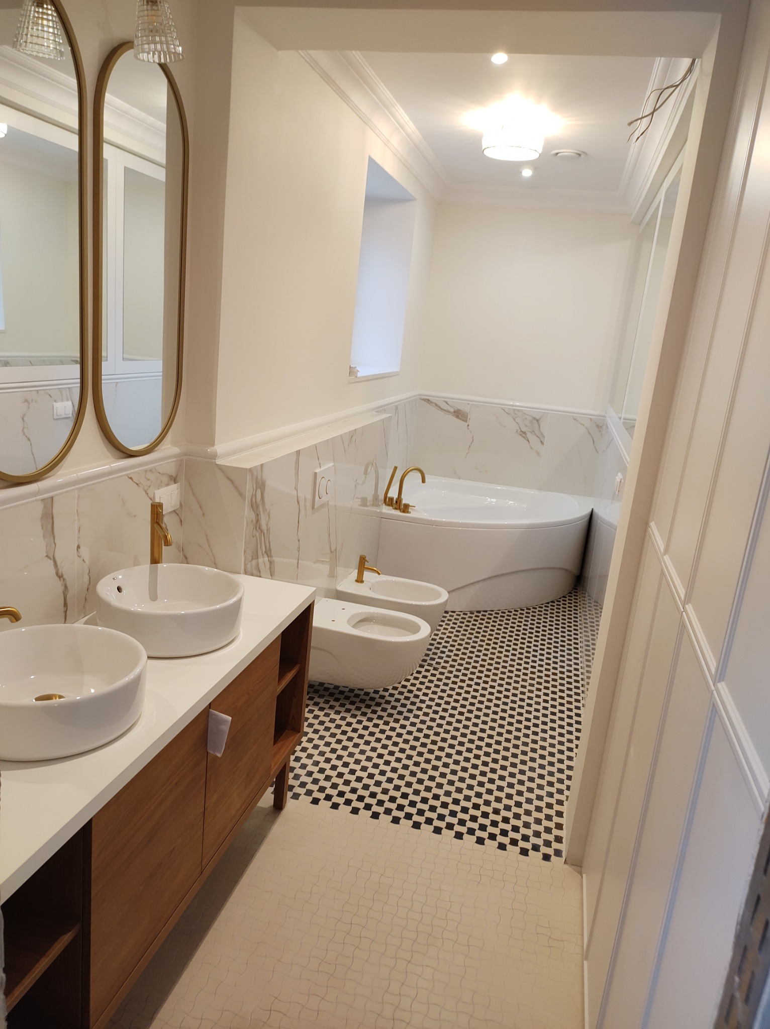 Nowoczesna łazienka w jasnych kolorach z marmurowymi akcentami, złotą armaturą, dwiema umywalkami na drewnianej szafce, bidetem, wanną oraz czarno-białą mozaiką na podłodze.