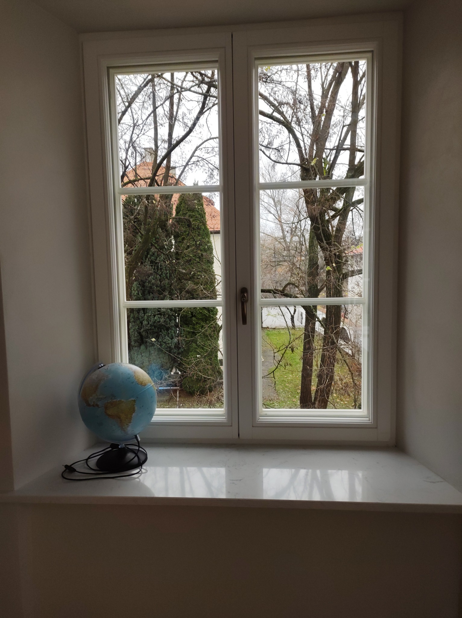 Dwuskrzydłowe okno z widokiem na ogród z drzewami bez liści, na parapecie globus z czarnym kablem.