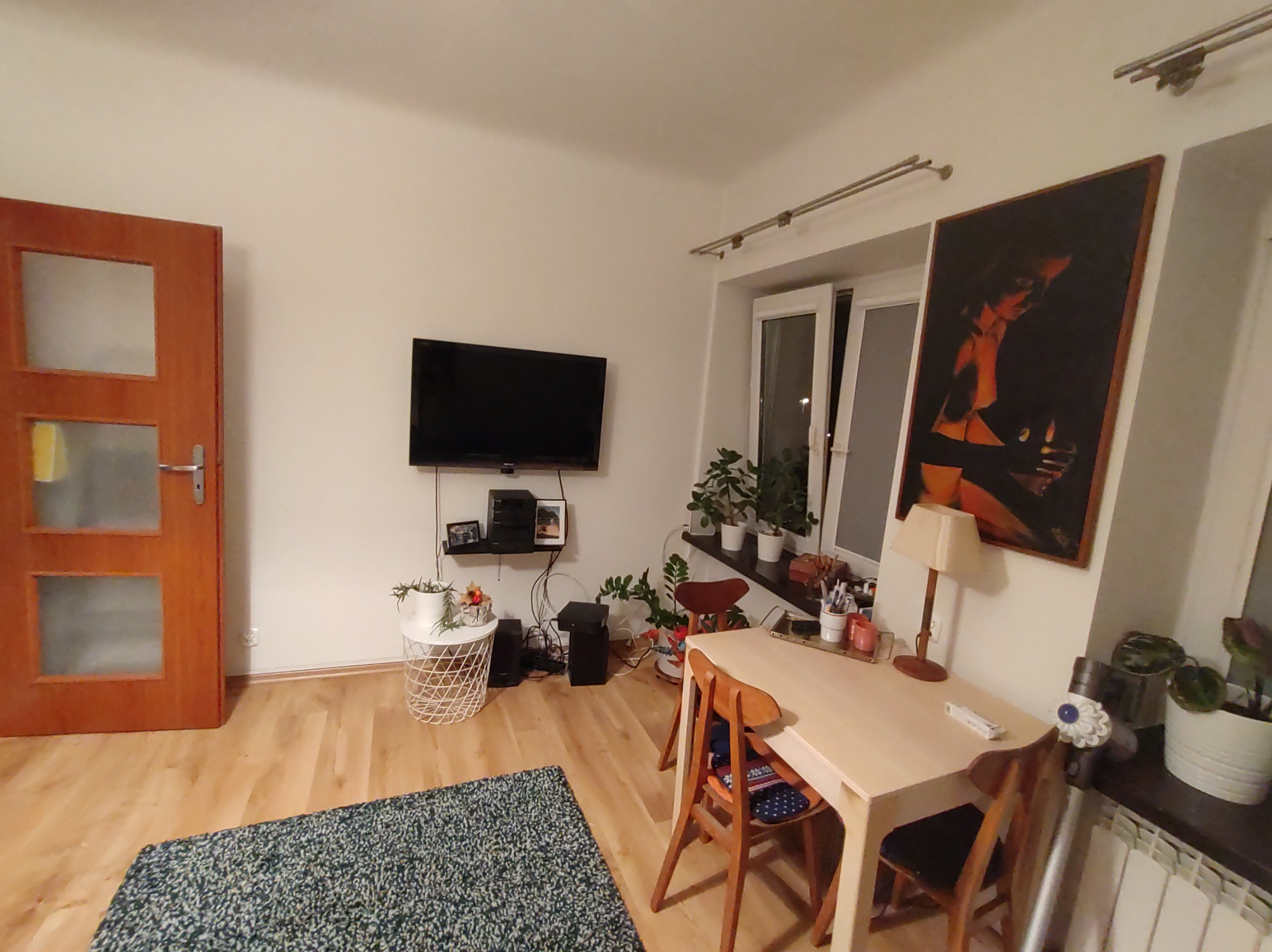 Widok na umeblowany pokój dzienny z drewnianą podłogą, dywanem w odcieniach zieleni, stołem z krzesłami, telewizorem na ścianie i oknem z roślinami, po remoncie.