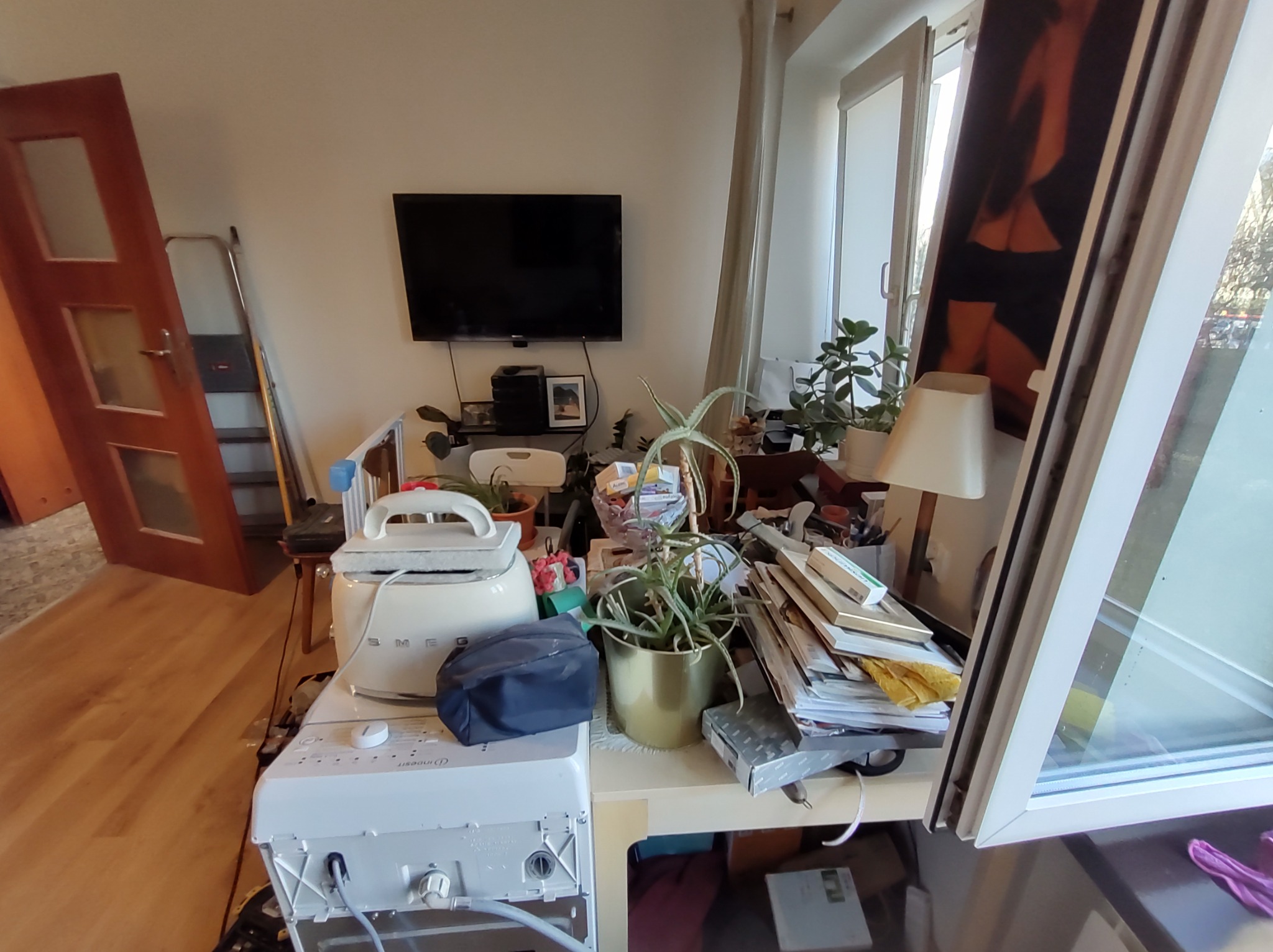 Zagracony pokój z widoczną deską do prasowania, żelazkiem parowym, stertą dokumentów, roślinami doniczkowymi i oknem. Widoczny bałagan na stole.