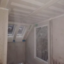 Poddasze w trakcie wykańczania: widoczne płyty na suficie i skośnych ścianach, przygotowane do dalszych prac wykończeniowych, okna dachowe i standardowe okno PCV.