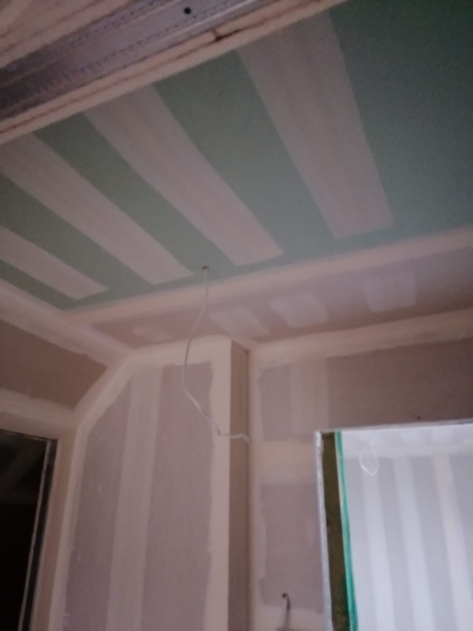 Wnętrze w trakcie remontu: widok na sufit z zamontowaną metalową konstrukcją i częściowo przytwierdzonymi płytami, w tym zielonymi płytami ognioodpornymi, ściany z widocznymi łączeniami płyt...