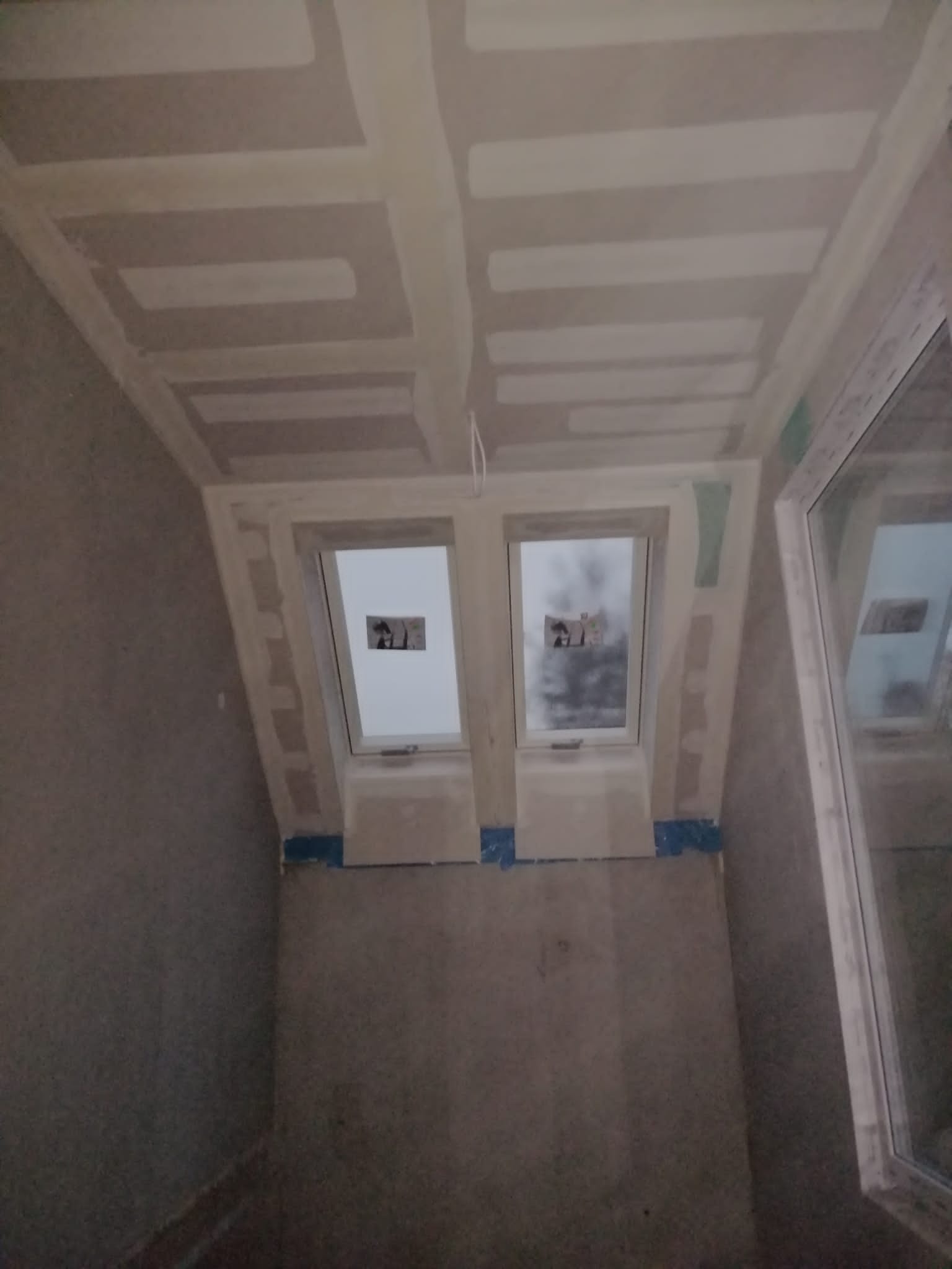 Poddasze w trakcie wykańczania: widoczne dwa okna dachowe, fragment okna pionowego po prawej, ściany i sufit pokryte płytami gipsowo-kartonowymi, na łączeniach widoczne szpachlowanie, taśma...