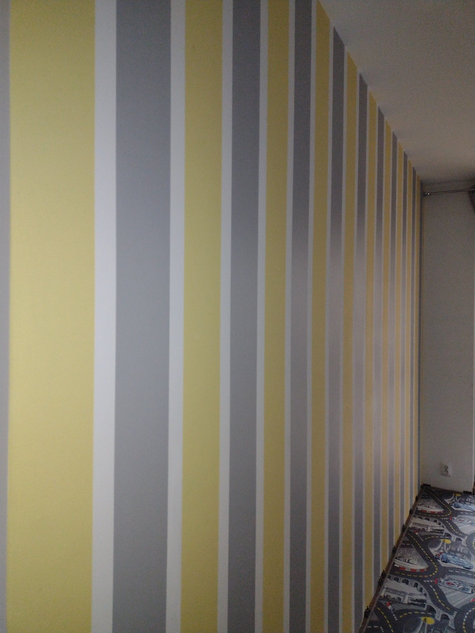 Pionowe pasy w kolorach szarym, żółtym i białym na ścianie, widok z dołu, fragment podłogi z wzorem toru wyścigowego.