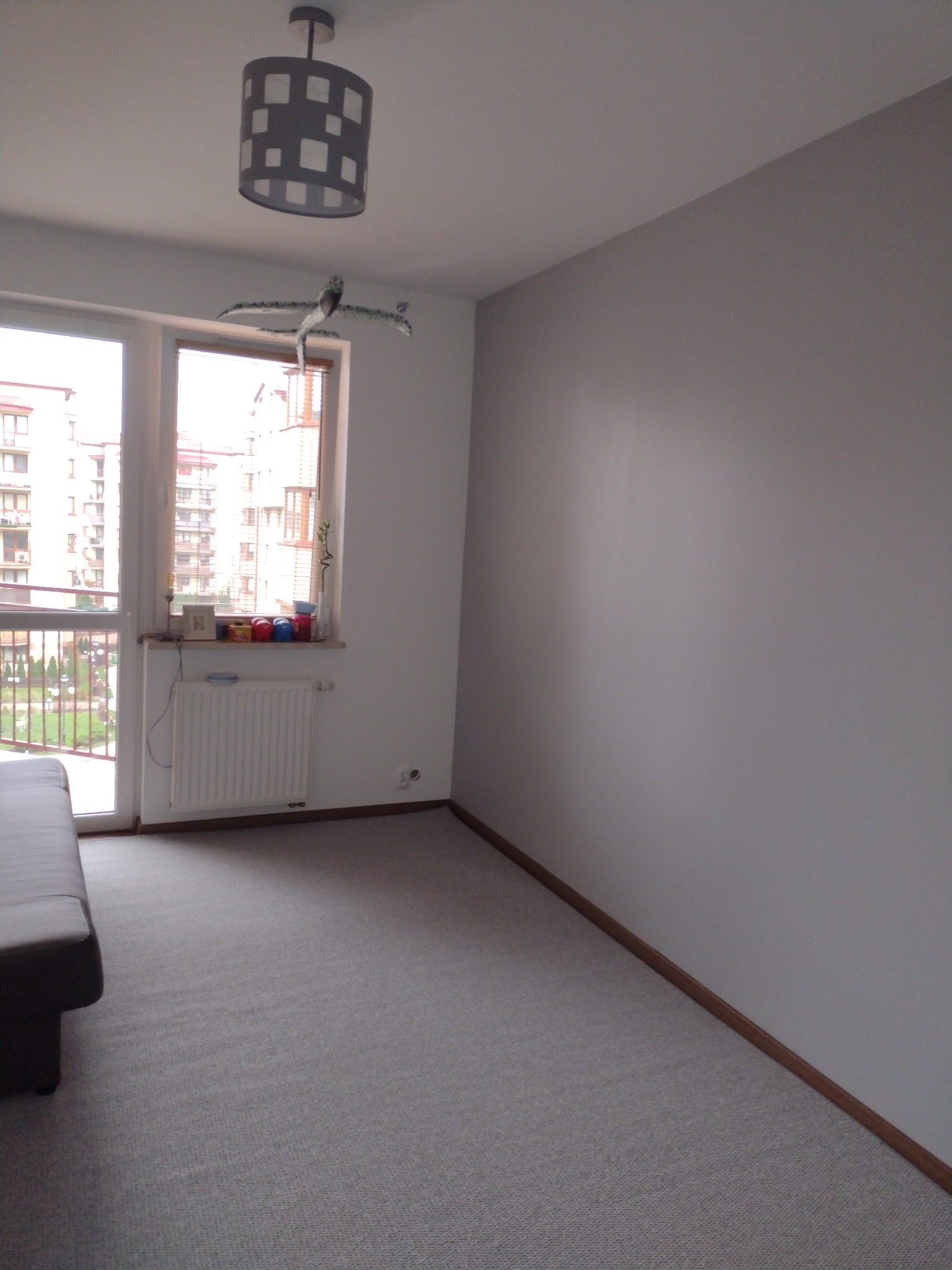 Świeżo pomalowane na szaro wnętrze pokoju w mieszkaniu w Warszawie, z widocznym oknem i grzejnikiem, szarą podłogą i ozdobną lampą sufitową.