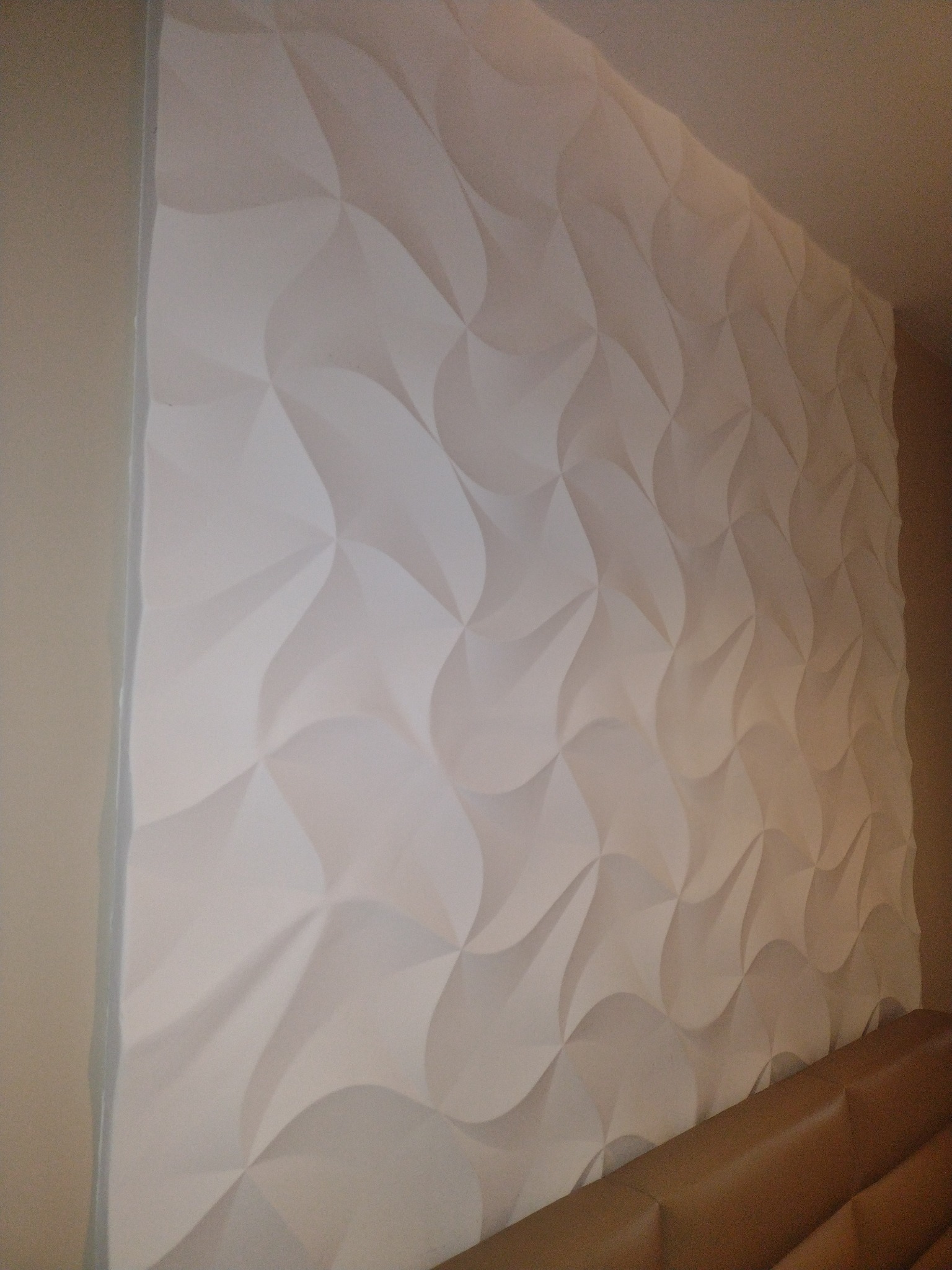 Ściana z dekoracyjnymi, trójwymiarowymi panelami gipsowymi o wzorze falistym, pomalowana na biało, widoczna z boku, z fragmentem brązowego zagłówka łóżka w dolnej części kadru.