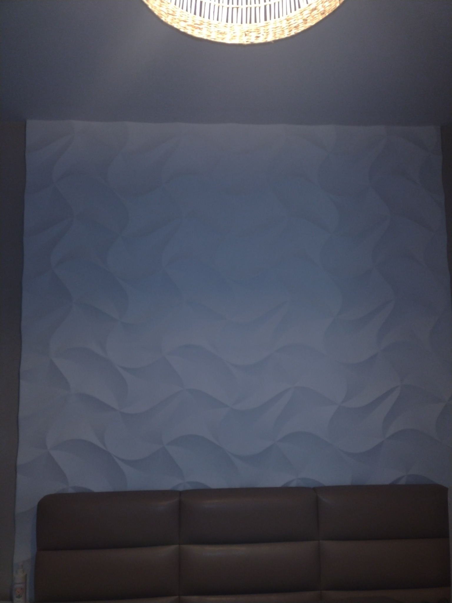 Ściana z dekoracyjnymi panelami 3D w odcieniach szarości, oświetlona od góry lampą z wikliny, z widocznym fragmentem wezgłowia łóżka.