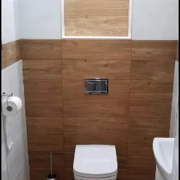 Wykończona łazienka z drewnopodobnymi płytkami na ścianach i podłodze, wisząca toaleta, mała umywalka i srebrny uchwyt na papier toaletowy.