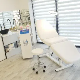 Jasne wnętrze gabinetu kosmetycznego z białym fotelem zabiegowym, taboretem, wózkiem z kosmetykami i szafkami z umywalkami na tle lustrzanej ściany i okna z żaluzjami.