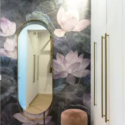 Przedpokój z tapetą w motywie kwiatów lotosu, owalne lustro w złotej ramie odbija wnętrze garderoby, pufa welurowa w kolorze pudrowego różu z podstawą w kolorze złota, biała szafa z podłużnymi...