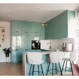 Nowoczesna kuchnia z błyszczącymi, miętowo-zielonymi szafkami, białym blatem, złotą armaturą i trzema welurowymi hokerami na tle wzorzystej zasłony.