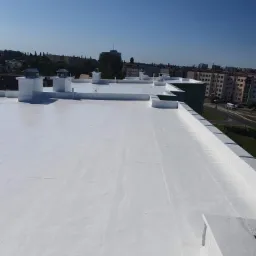 Biała, płaska powierzchnia dachu po renowacji, widok z góry na tle zabudowy miejskiej w słoneczny dzień.