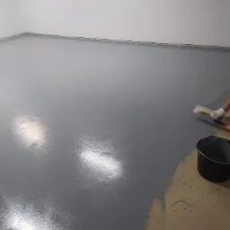 Aplikacja szarej, błyszczącej powłoki na podłodze w pomieszczeniu, widoczny pracownik w rękawicy rozprowadzający materiał packą oraz czarne wiadro z pozostałościami.