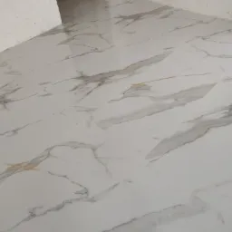 Błyszcząca podłoga wyłożona białymi płytkami imitującymi marmur z szarymi i złotymi żyłami, widoczne niedokończone ściany i gniazdko elektryczne.