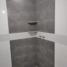 Wnętrze łazienki w trakcie prac wykończeniowych: szare płytki imitujące beton na ścianach, białe płytki na górze i po bokach, półki wnękowe, wystające rury ciepłej i zimnej wody, widoczne...