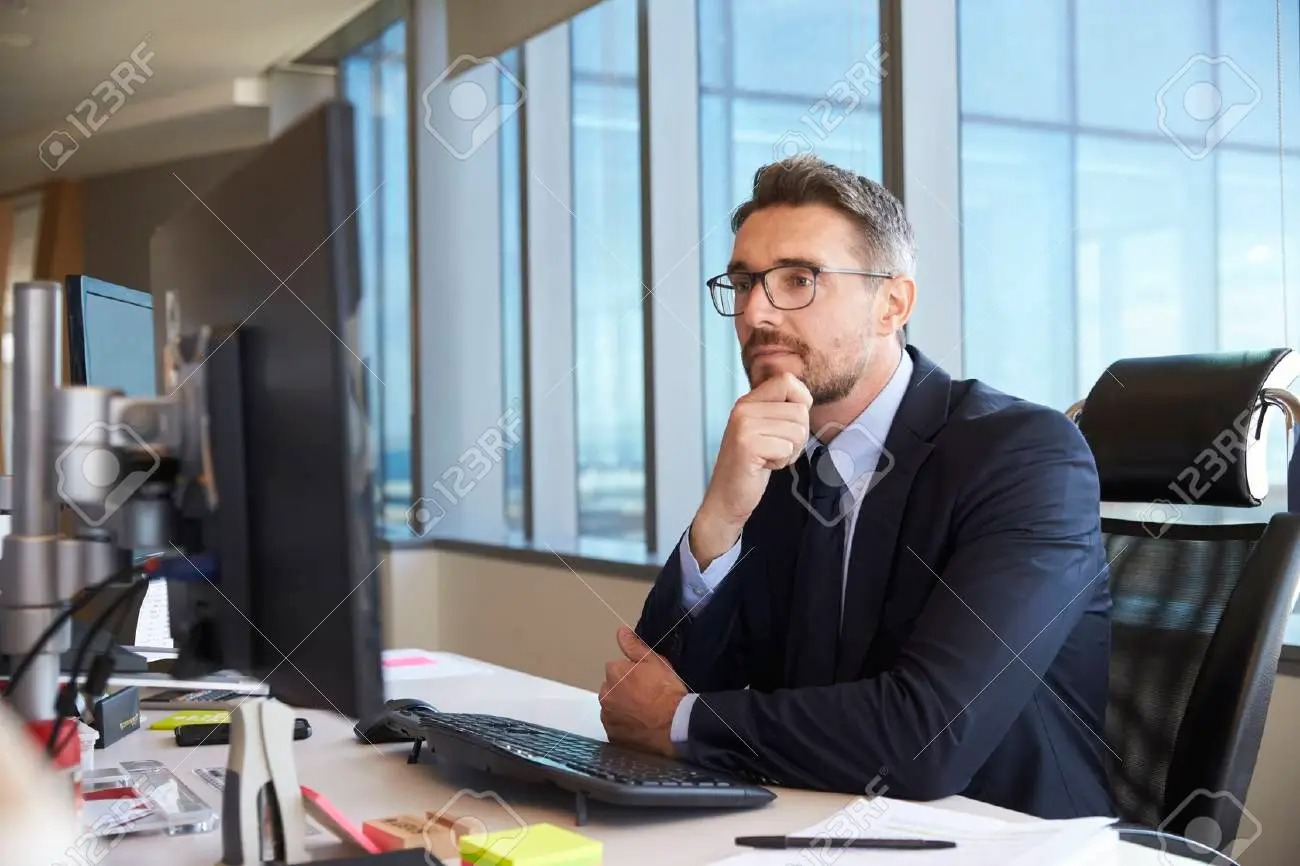 Mężczyzna w garniturze i okularach siedzi przy biurku z komputerem, zamyślony, z ręką podpartą pod brodą, w tle okno z widokiem na miasto.