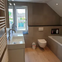 Nowoczesna łazienka na poddaszu z drewnianą podłogą, dwoma umywalkami nablatowymi, podwieszaną toaletą i wanną w zabudowie, oświetlona punktowymi lampami.