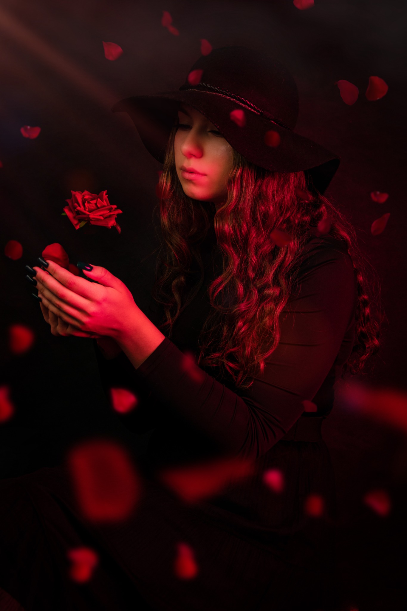 Portret kobiety w czarnym kapeluszu, trzymającej czerwoną różę w dłoniach z ciemnozielonymi paznokciami, otoczonej opadającymi płatkami róż w czerwonym oświetleniu.