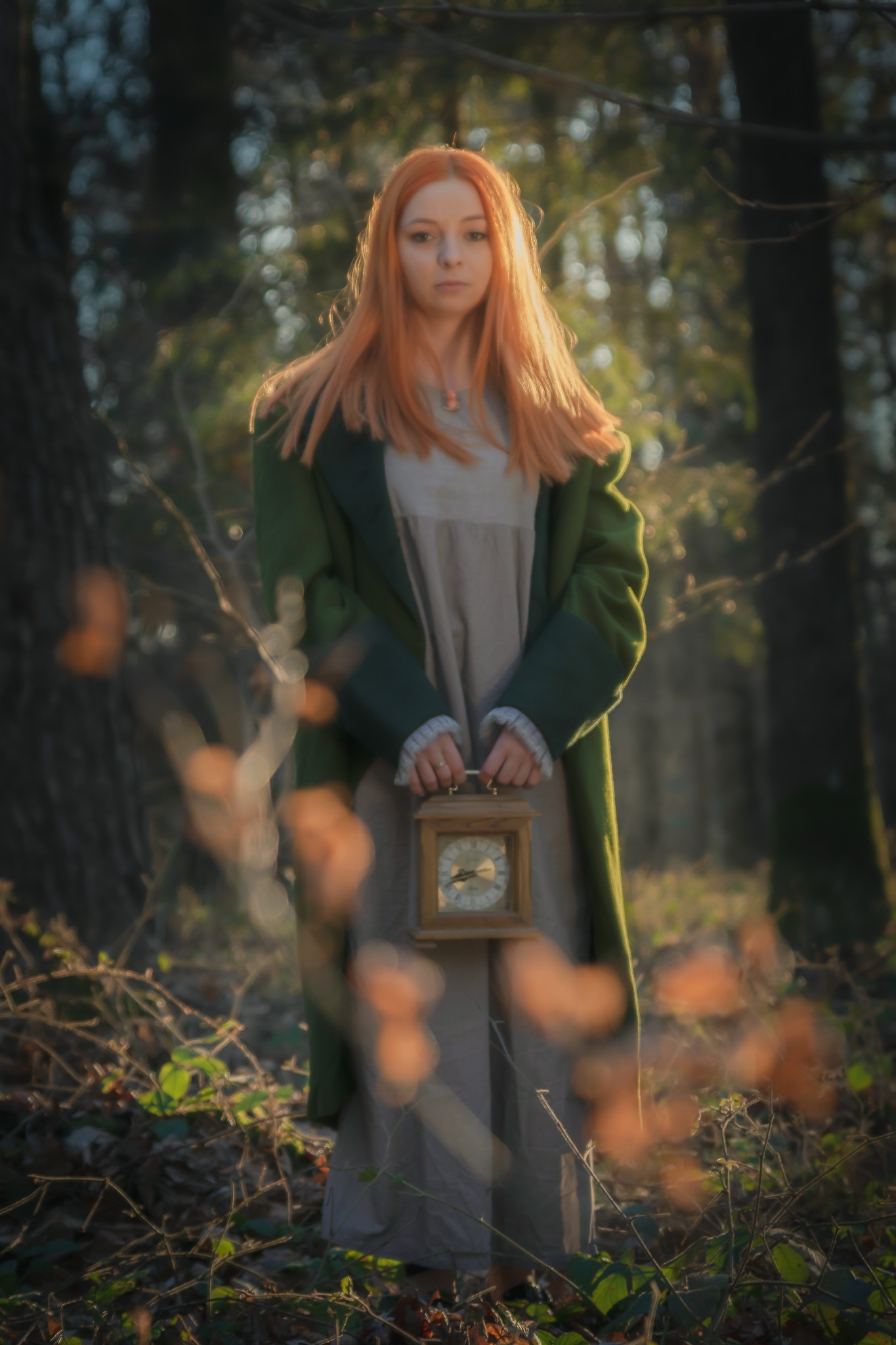 Rudowłosa kobieta w zielonym płaszczu trzymająca stary zegar w drewnianej obudowie, stojąca w zalesionym otoczeniu z rozproszonym światłem.