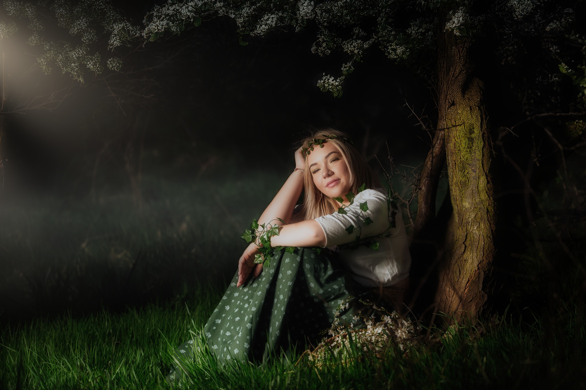 Kobieca sesja w Woli Radziszowskiej: Blondwłosa kobieta w wianku z bluszczu, siedząca pod kwitnącym drzewem w leśnej scenerii, w zielonej sukience i białej bluzce.