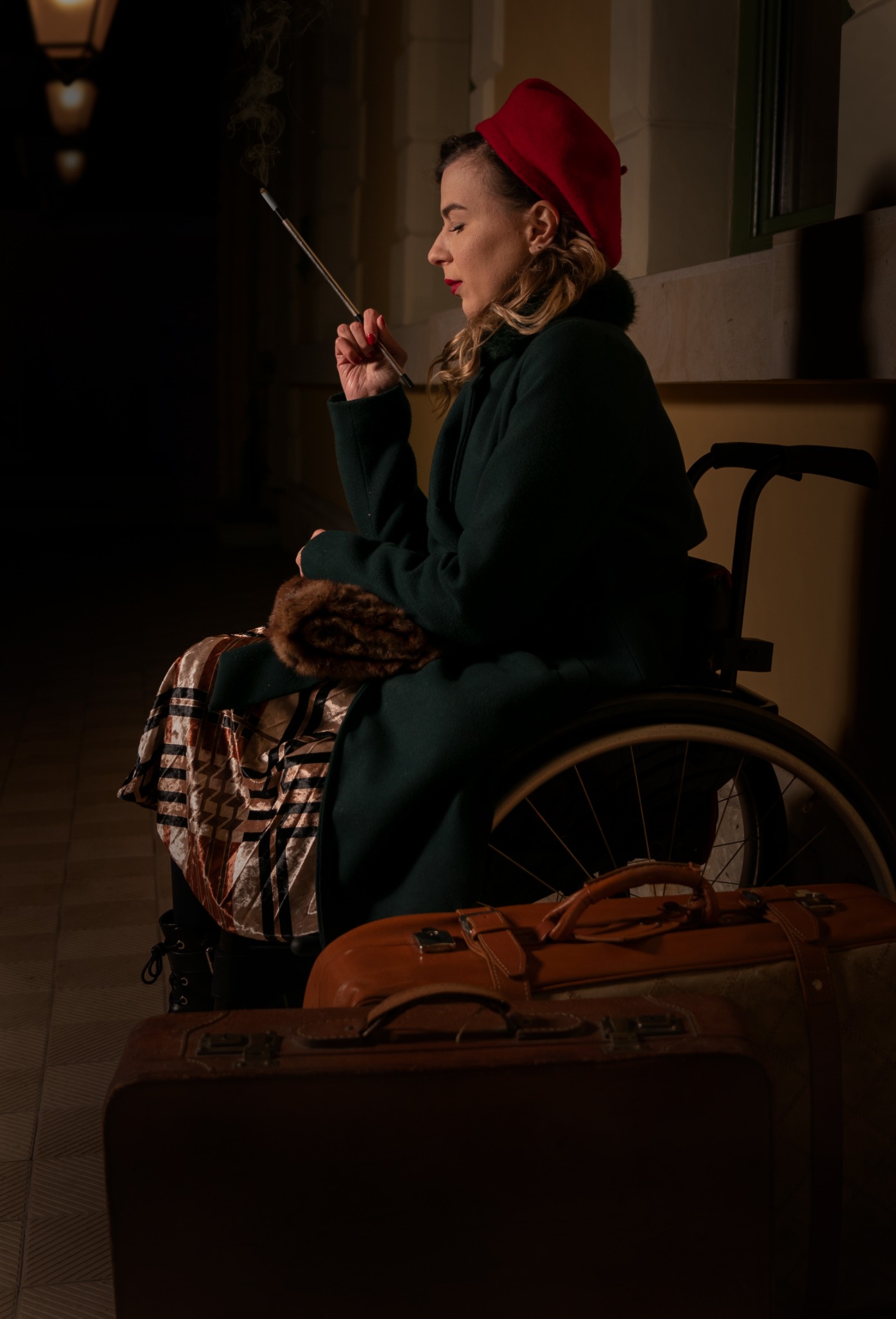 Kobieta w czerwonym berecie i zielonym płaszczu, siedząca na wózku inwalidzkim, pali papierosa w lufce na tle walizek i oświetlonego budynku.