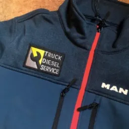 Granatowo-błękitna bluza z logo firmy Truck Diesel Service na lewej piersi i napisem MAN na dole po prawej stronie, zapinana na zamek z czerwonym paskiem pośrodku.