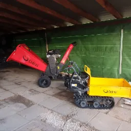 Czerwony rozdrabniacz do gałęzi i ż&oacute;łta mini wywrotka gąsienicowa marki Cedrus stoją na betonowej nawierzchni pod zadaszeniem. W tle zielona plandeka.