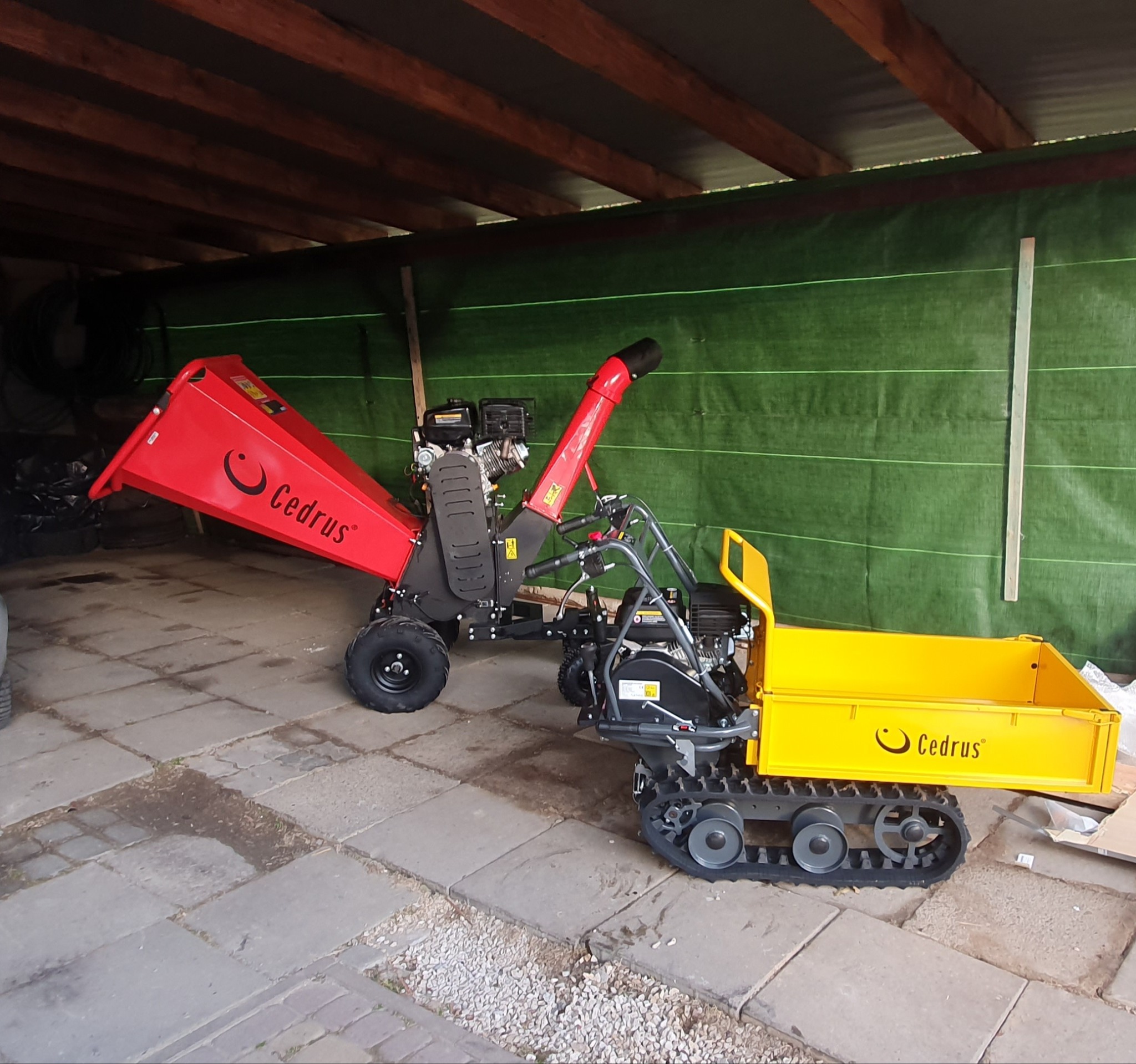 Czerwony rozdrabniacz do gałęzi i żółta mini wywrotka gąsienicowa marki Cedrus stoją na betonowej nawierzchni pod zadaszeniem. W tle zielona plandeka.