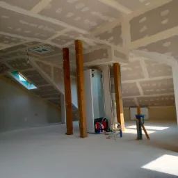 Poddasze w trakcie wykańczania: ściany i sufit pokryte płytami, z widocznymi łączeniami przed szpachlowaniem, drewniane podpory, okno dachowe i instalacje w tle.