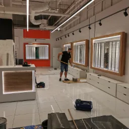 Mężczyzna z brodą rozprowadza gładź szpachlową na podłodze w nowym lokalu optycznym, widoczne narzędzia malarskie, białe płytki podłogowe i meble w tle.