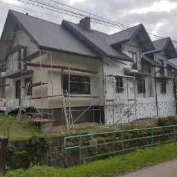 Dwukondygnacyjny dom jednorodzinny z ciemnym dachem w trakcie prac elewacyjnych, widoczne rusztowania i częściowo ocieplona ściana z białym styropianem, tło stanowią drzewa i pochmurne niebo.