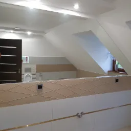 Ukończona łazienka na poddaszu z białymi i beżowymi płytkami, dekoracyjnym wzorem z cienkich listew, widoczne elementy armatury i toaleta, odbicie w lustrze.