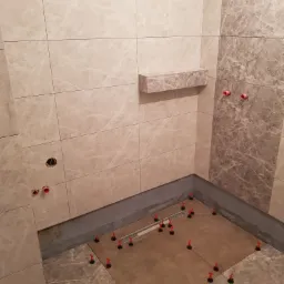 Układanie płytek w kabinie prysznicowej z odpływem liniowym, użycie czerwonych klipsów poziomujących, marmurowe płytki na ścianach i podłodze, półka wnękowa.