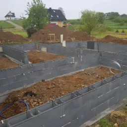 Fundamenty z szarych bloczków betonowych, zbrojenie pionowe, przygotowane pod wylewkę, w tle dom w trakcie budowy i koparka.