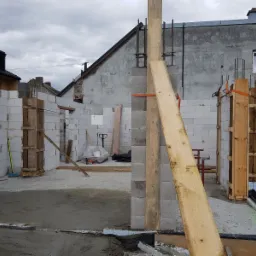 Wznoszenie ścian z bloczków betonowych na fundamencie, widoczne drewniane szalunki kolumn, zbrojenia pionowe i narzędzia budowlane w tle.