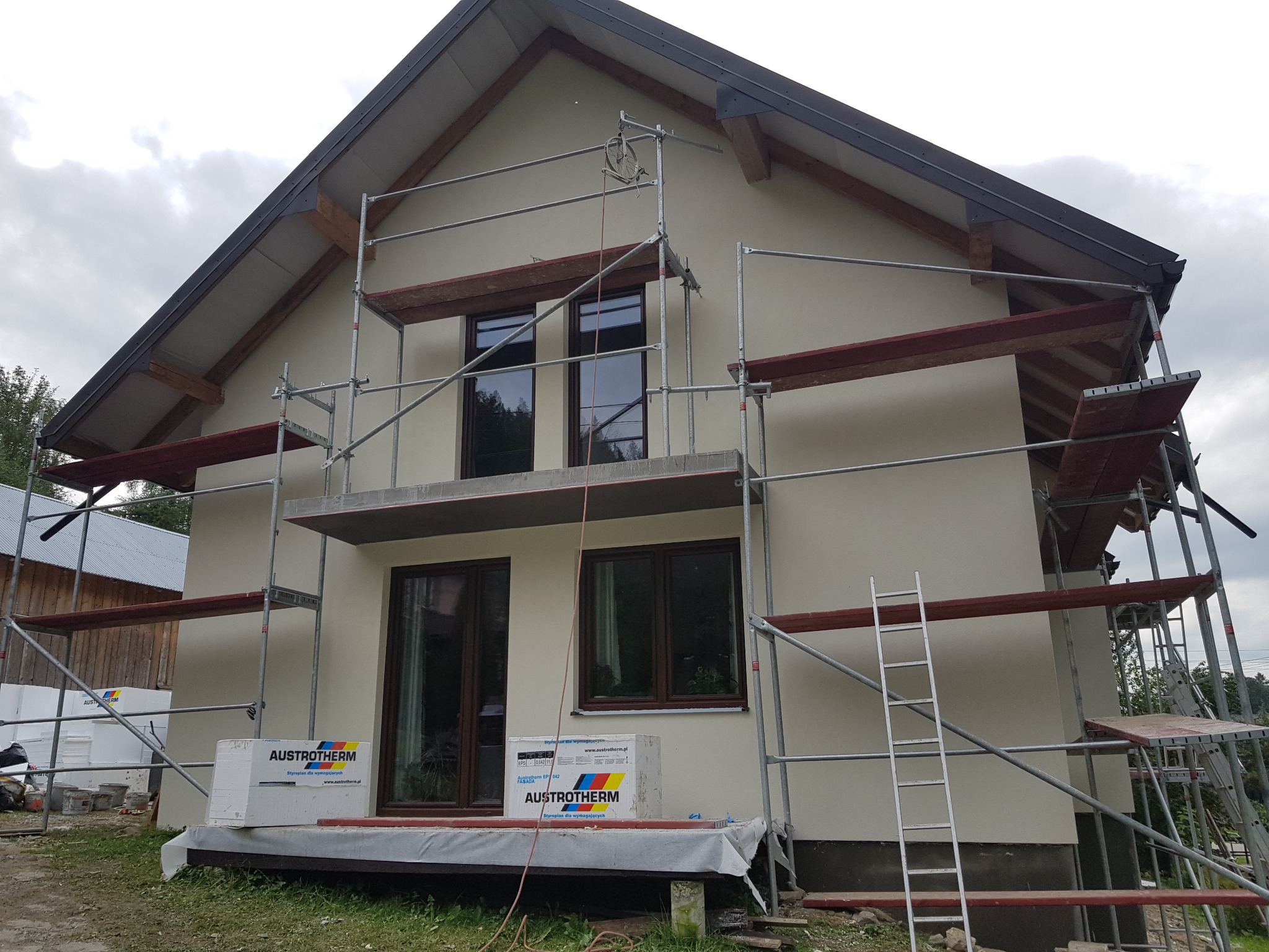 Dwukondygnacyjny dom w trakcie ocieplania elewacji, widoczne rusztowania, drabina oraz paczki styropianu Austrotherm, częściowo otynkowana ściana w kolorze beżowym, brązowe ramy okienne.