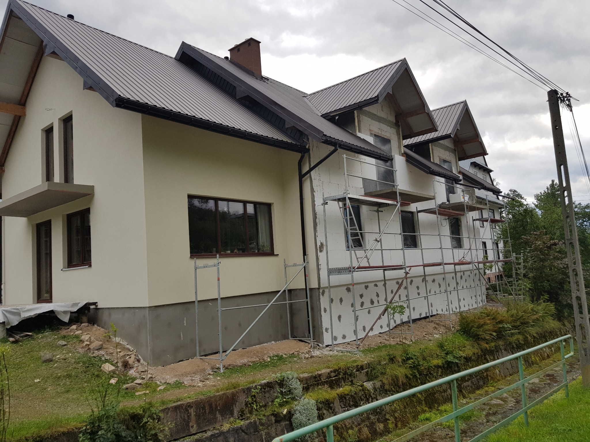 Dwukondygnacyjny dom z szarą metalową blachodachówką, częściowo ocieplony styropianem, z widocznymi rusztowaniami i fragmentem elewacji w kolorze kremowym.