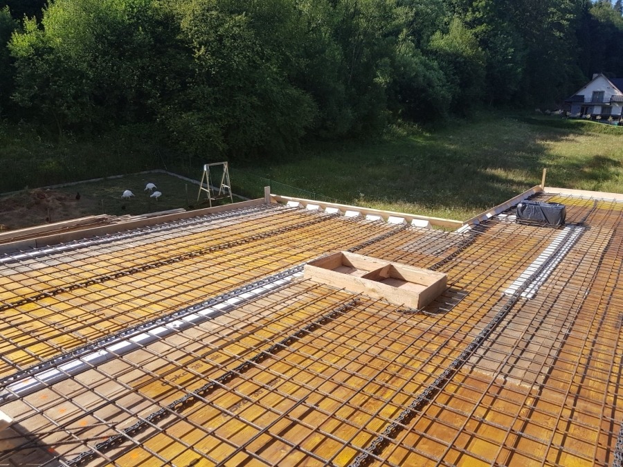 Widok z góry na szalunek stropu z siatką zbrojeniową i elementami drewnianymi, w tle trawnik, drzewa i dom.