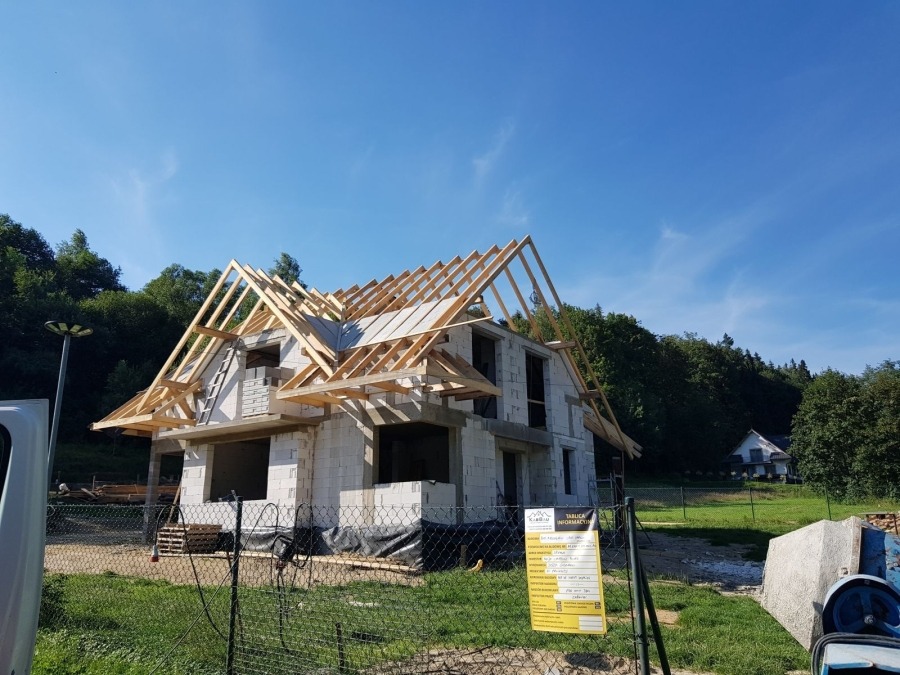 Dwukondygnacyjny dom w trakcie budowy, z widoczną konstrukcją drewnianą dachu i ścianami z białych bloczków, na tle zieleni i błękitnego nieba. Tablica informacyjna budowy widoczna z przodu.