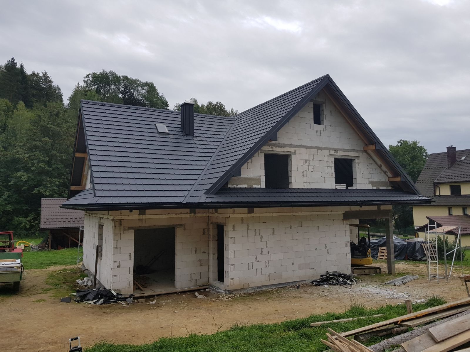 Niedokończony dom z białego bloczka z zamontowanym czarnym dachem z blachodachówki i kominem, widoczne otwory okienne bez okien, z tyłu drzewa.