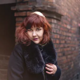 Portret kobiety w czarnym płaszczu z futrzanym kołnierzem na tle ceglanej ściany w Nikiszowcu. Uwagę zwraca jej ciemna szminka i oryginalne upięcie włosów.