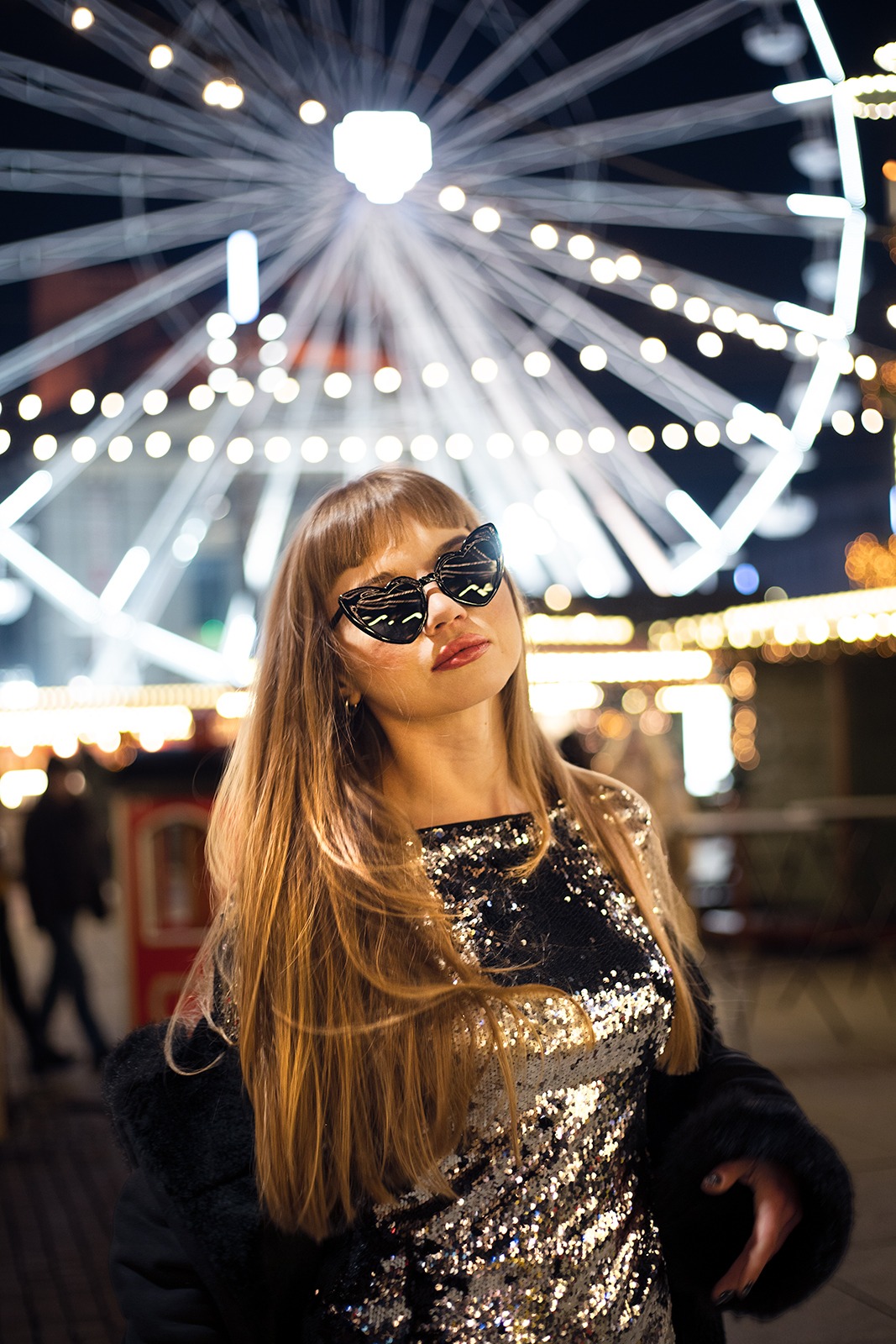 Portret kobiety w cekinowej sukience i okularach w kształcie serc na tle oświetlonego diabelskiego młyna nocą.