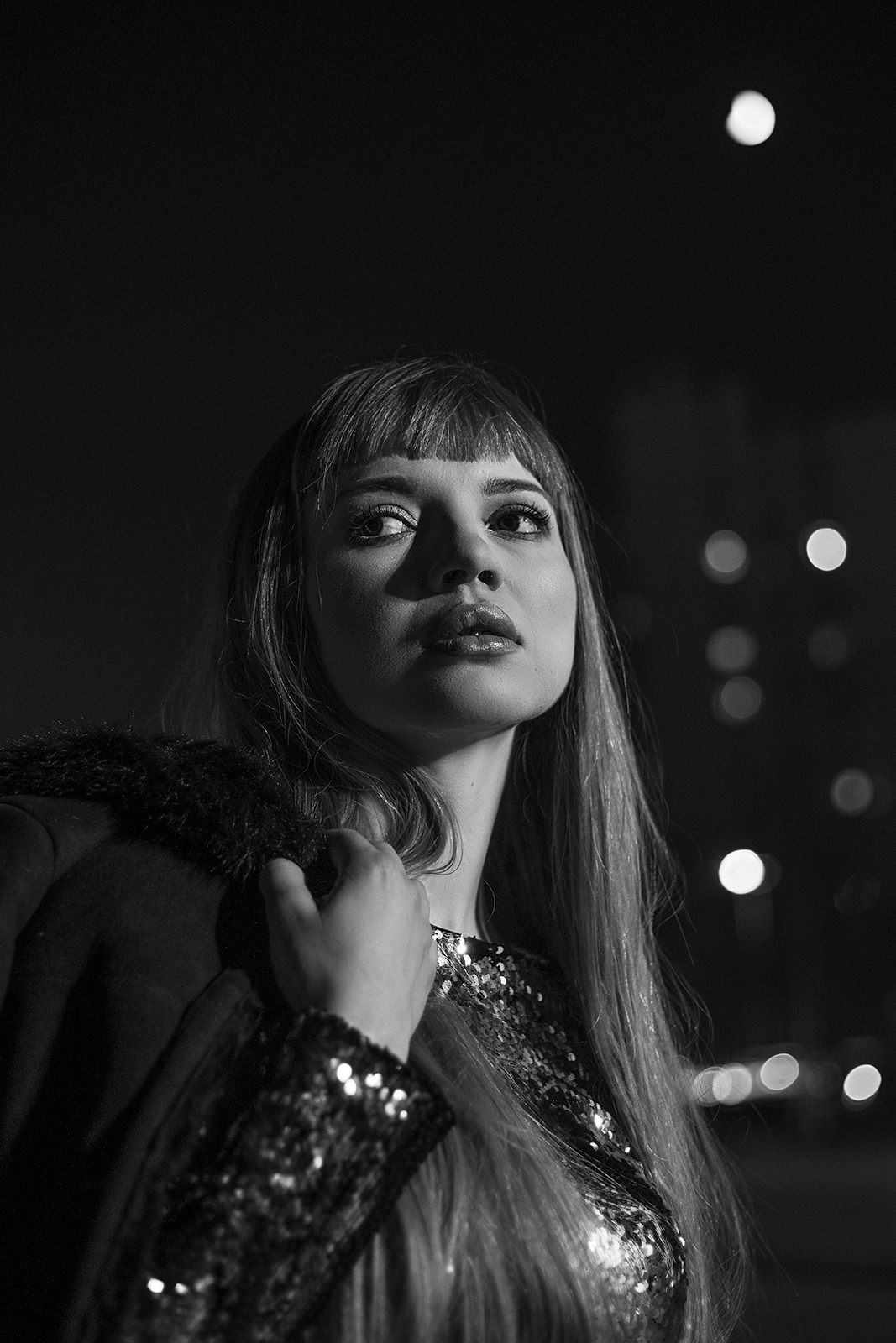 Portret kobiety w czarno-białej tonacji, spoglądającej w górę, z błyszczącą bluzką i futrzanym kołnierzem, na rozmytym tle świateł miejskich.