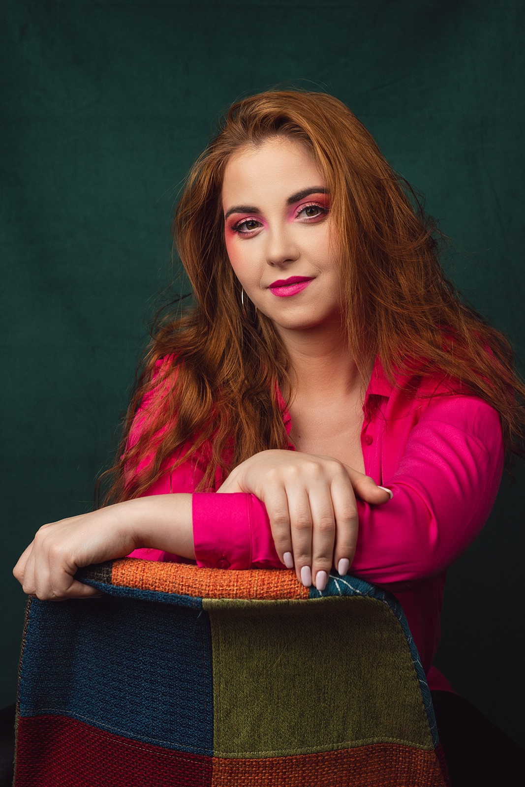 Portret kobiety z rudymi włosami i różowym makijażem, ubrana w różową koszulę, opierająca ręce o patchworkowe oparcie krzesła na tle ciemnozielonego tła.