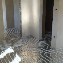 Białe rury ogrzewania podłogowego ułożone na folii w surowym wnętrzu z widocznymi otworami drzwiowymi w stanie deweloperskim.