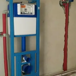 Stelaż podtynkowy WC w trakcie montażu, widoczne rury doprowadzające wodę i odpływ, instalacja ogrzewania podłogowego na podłodze, czerwone rury w ścianie.