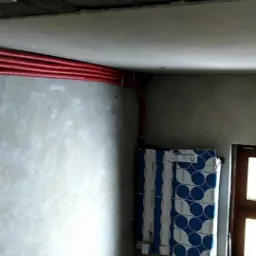 Wewnętrzny narożnik pomieszczenia w trakcie remontu; widoczne czerwone rury instalacji grzewczej poprowadzone pod sufitem, niezabezpieczony sufit z ubytkami, okno z drewnianą ramą oraz stos...